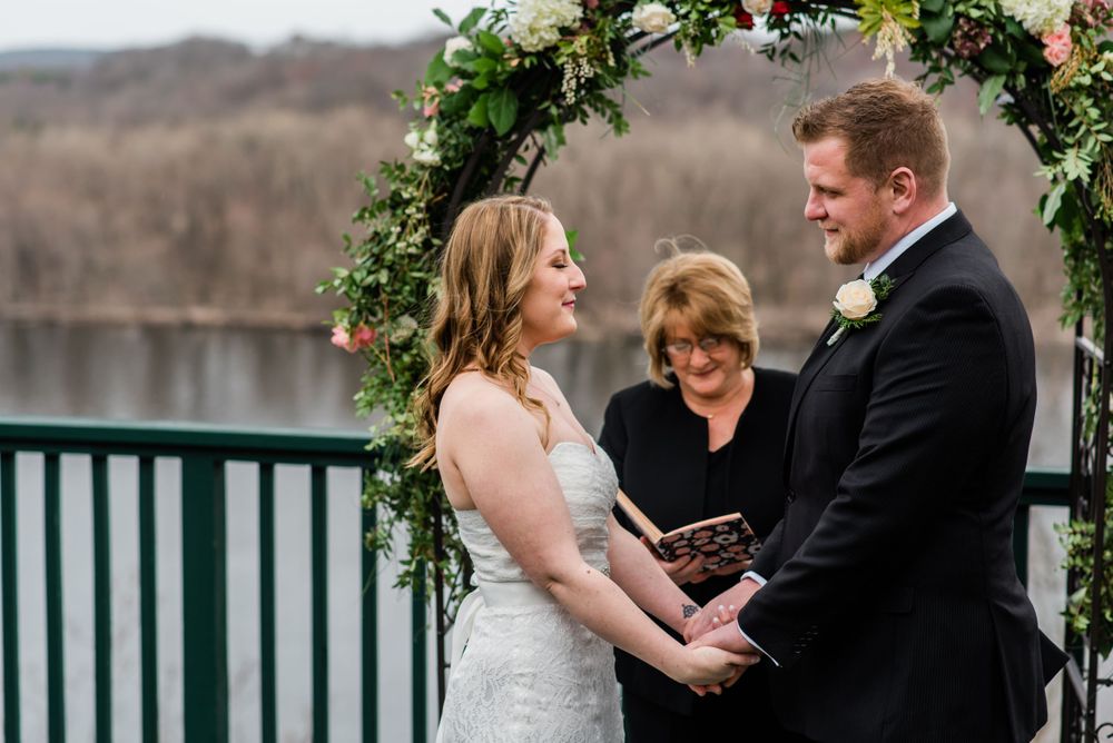 Wedding ceremony takes place under a floral arch on a lakeside deck during winter.