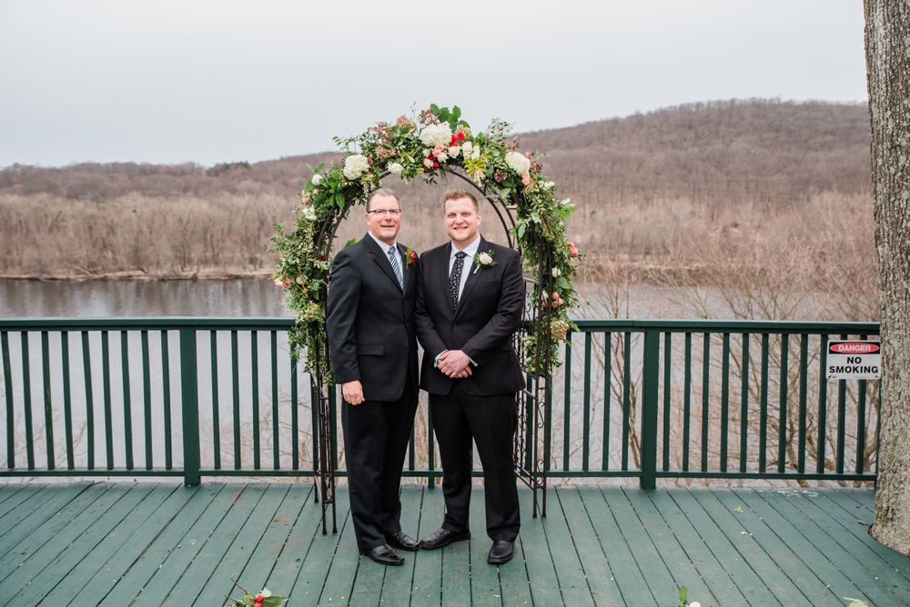 Two people stand under a floral arch on a green deck with a lake and winter landscape behind.