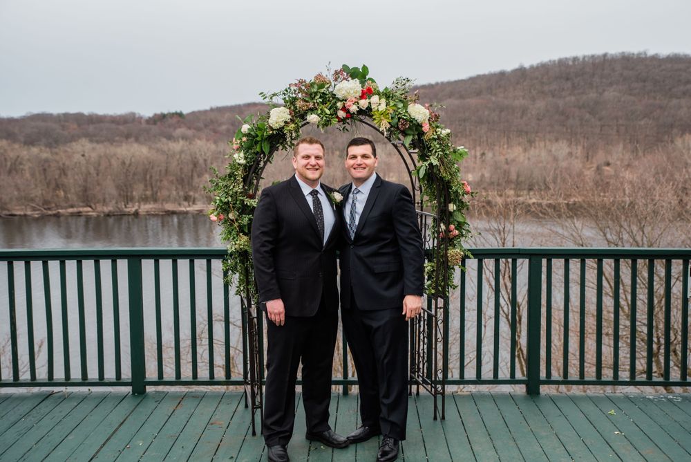 Two people in dark suits pose under a floral arch on a lakeside deck.