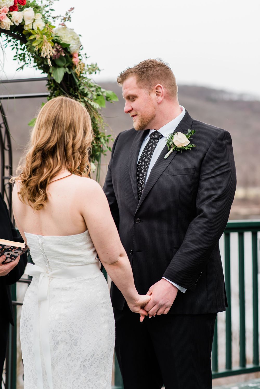 A wedding ceremony takes place under a floral arch on an outdoor deck overlooking a lake.