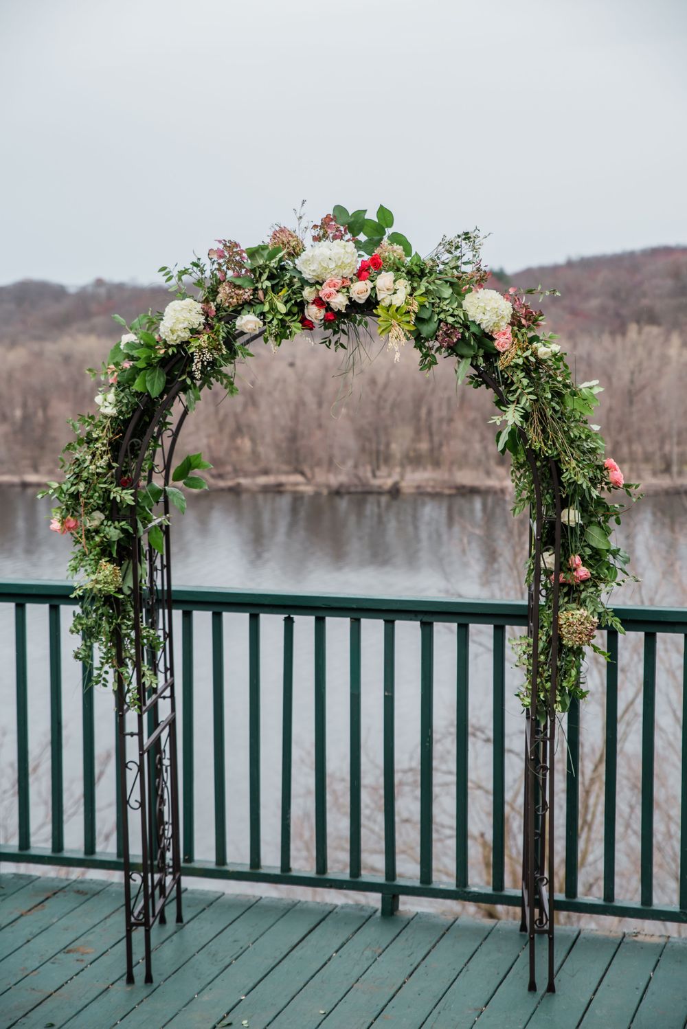 A metal wedding arch decorated with lush greenery and pink roses overlooks a serene lake.