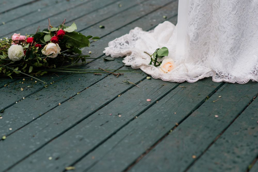 The hem of a white wedding dress trails on a teal deck next to scattered rose petals.