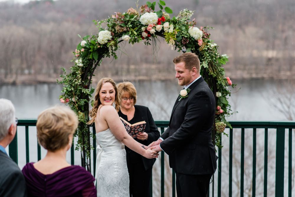 An intimate outdoor wedding ceremony beneath an ornate floral arch with water views.