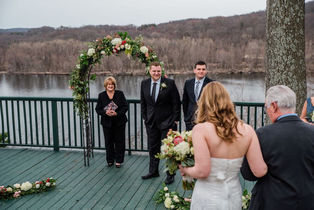 A wedding ceremony takes place on a lakeside deck under a floral arch with an officiant and guests.