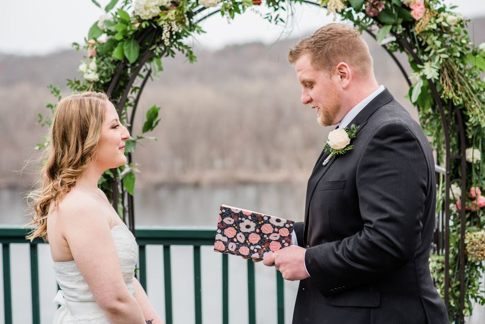 A couple exchanges vows beneath a floral arch on a deck overlooking a lake.