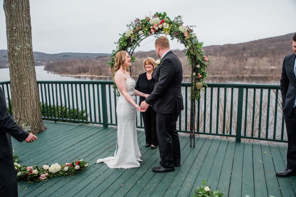 An intimate wedding ceremony takes place on a deck with a floral arch and lake backdrop.