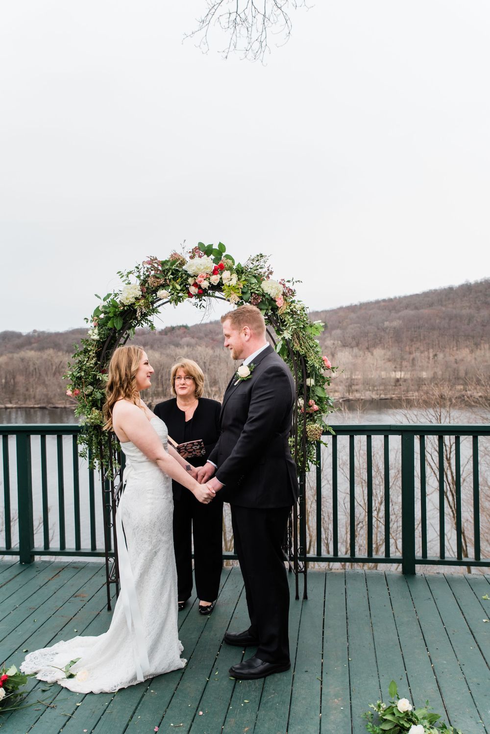 A wedding ceremony takes place on a deck overlooking winter hills under a floral arch with greenery and blooms.
