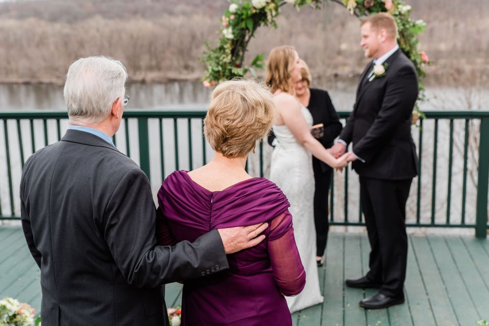 A wedding ceremony viewed from behind guests on an outdoor deck with a floral arch and scenic backdrop.