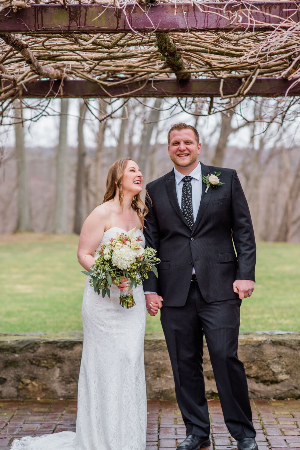 A joyful couple in wedding attire poses under a rustic branch archway on a brick path.