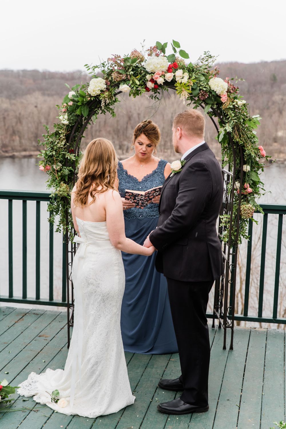 A wedding ceremony takes place under a floral arch on a deck overlooking a lake.