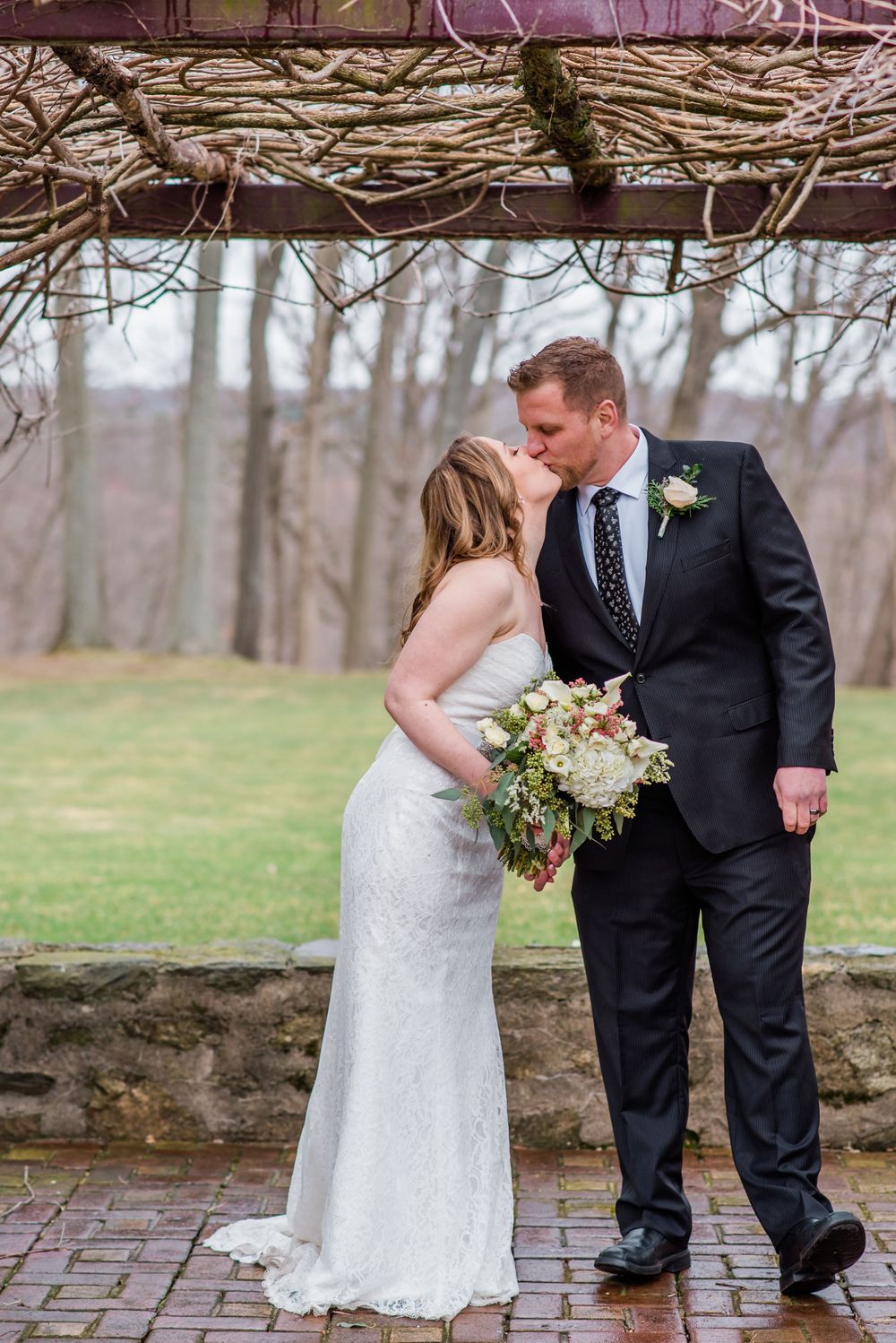 A couple in formal wedding attire share an intimate moment under a rustic arbor outdoors.