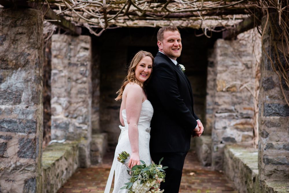 A couple poses in formal wedding attire in front of a stone archway.