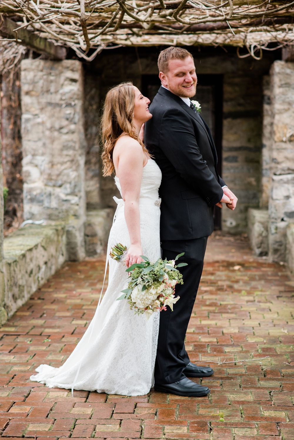 A couple poses back-to-back on brick pavement under a rustic branch covering.