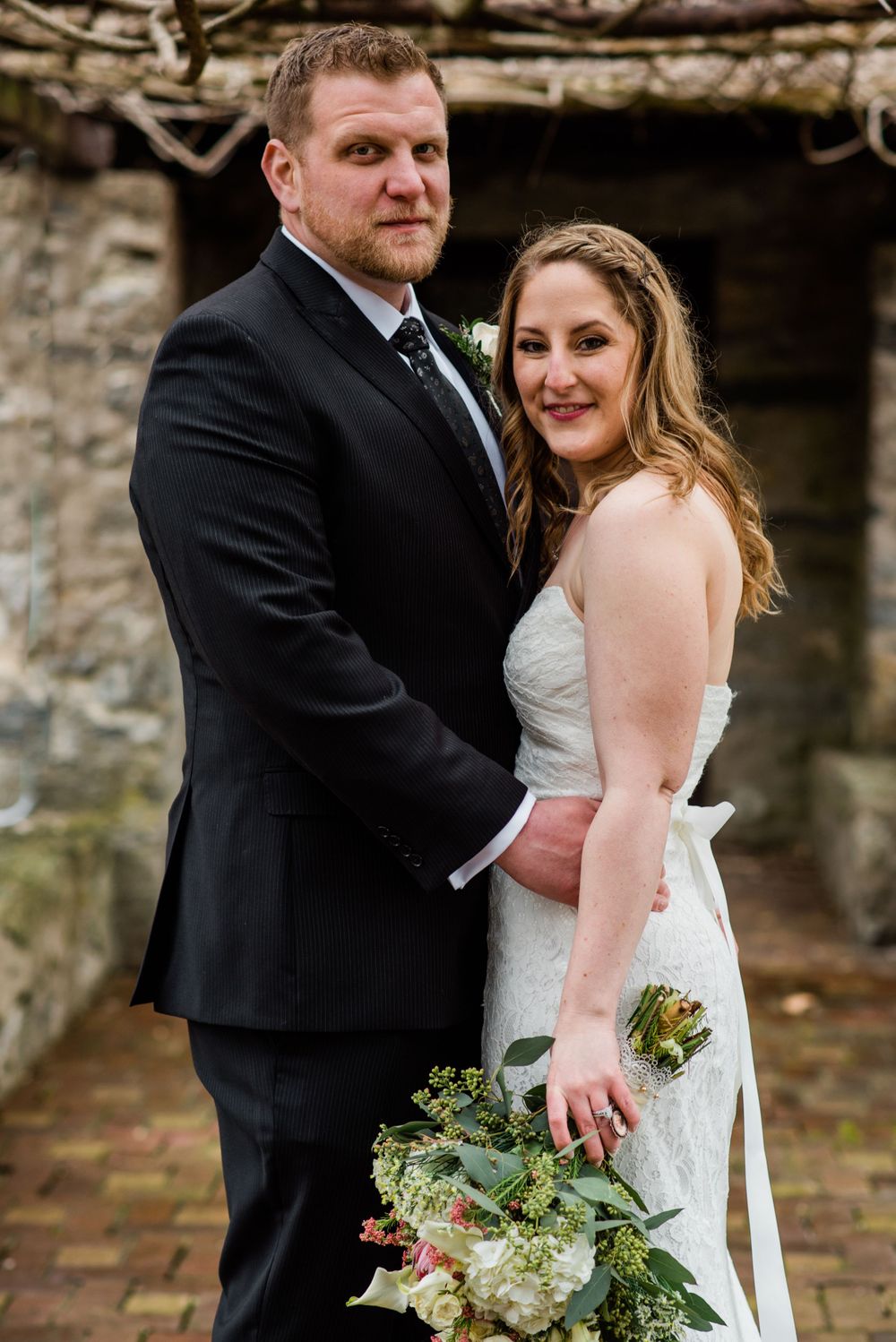 A couple embraces in wedding attire in front of a stone structure with brick flooring.