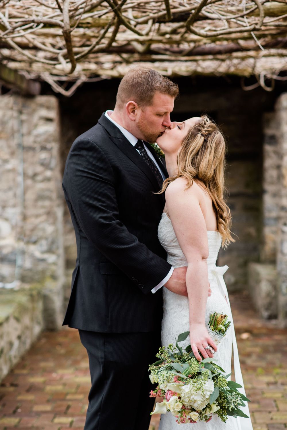 A couple shares an intimate kiss during their wedding photoshoot against a stone wall backdrop.