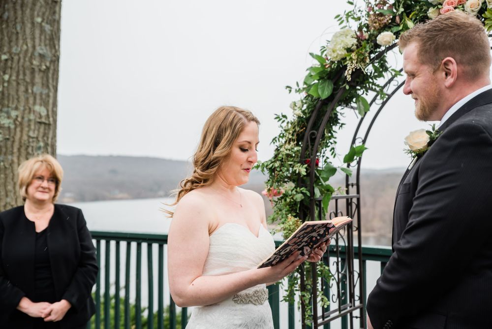 An outdoor wedding ceremony takes place under a floral arch with a waterfront view.