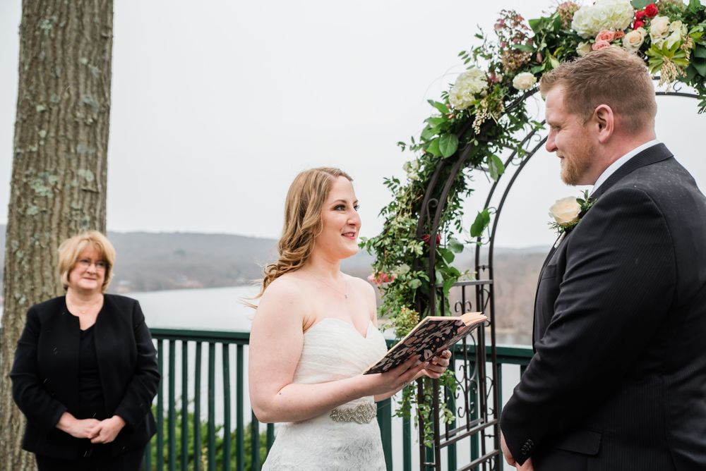 A bride reads vows during an outdoor ceremony beneath a decorated floral arch.