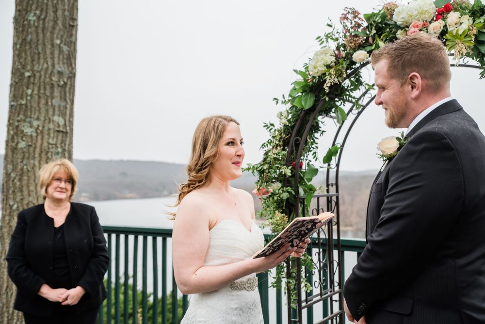 Outdoor wedding ceremony with couple exchanging vows under floral arch overlooking water.