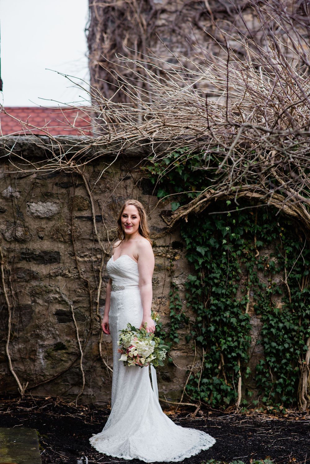 A bride in white dress poses against stone wall with ivy and bare branches.