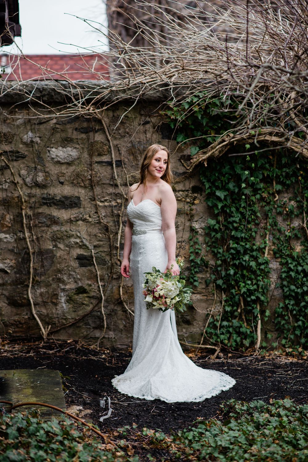 Bride in a fitted white lace gown poses against rustic stone wall with ivy backdrop.