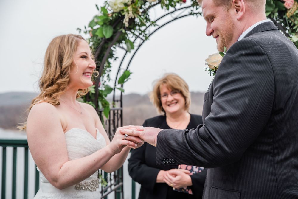 Wedding ceremony under floral arch with couple exchanging rings on outdoor deck.