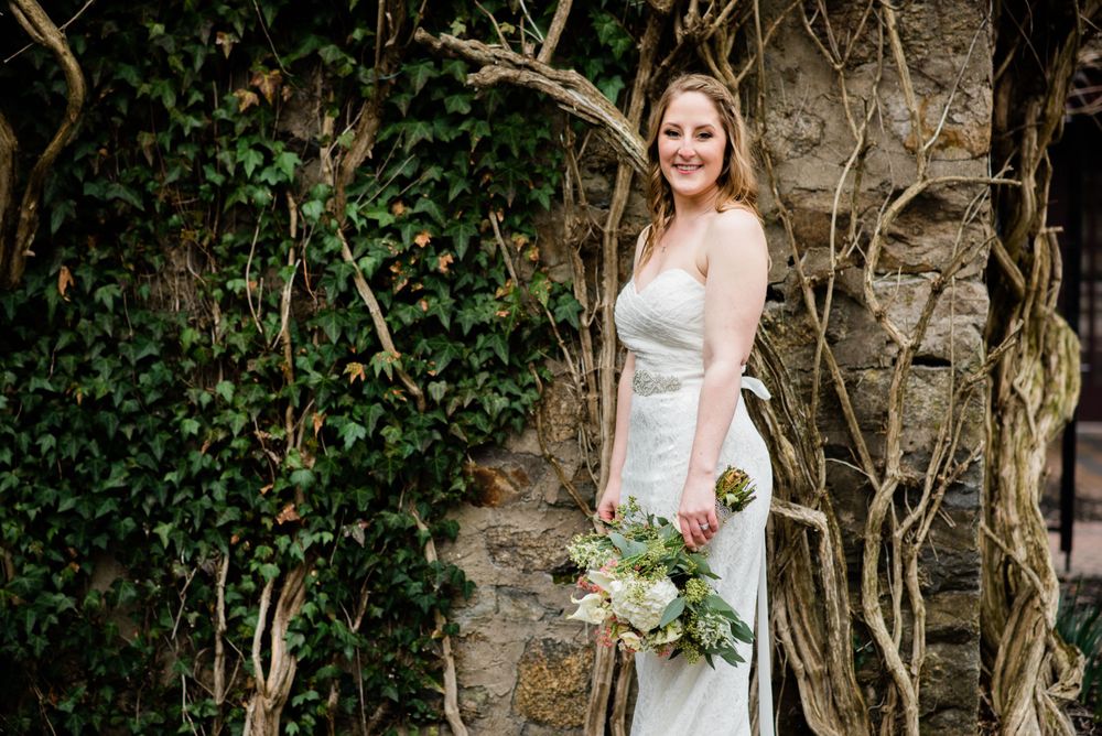 Elegant bride in fitted white dress poses against ivy-covered stone wall.