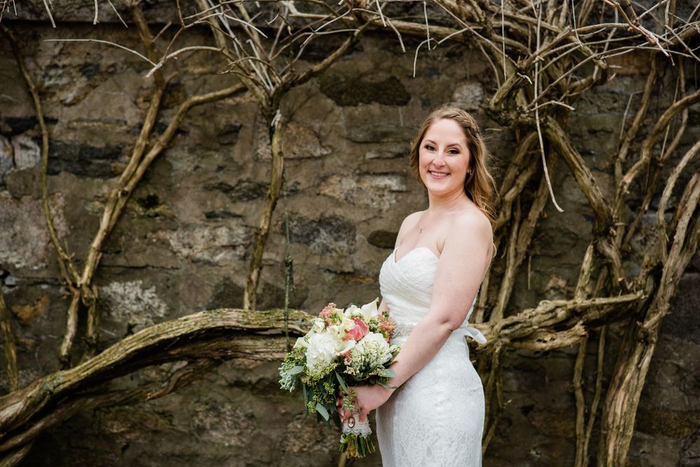 Bride holding bouquet poses against stone wall with bare vines.