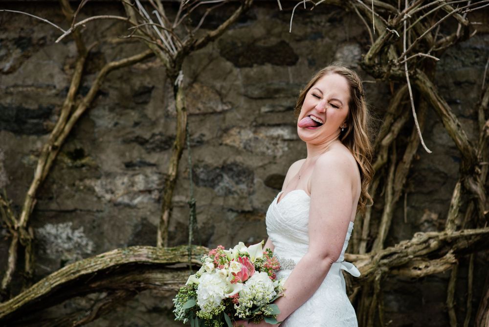 Joyful bride laughs while holding bouquet against rustic stone backdrop.