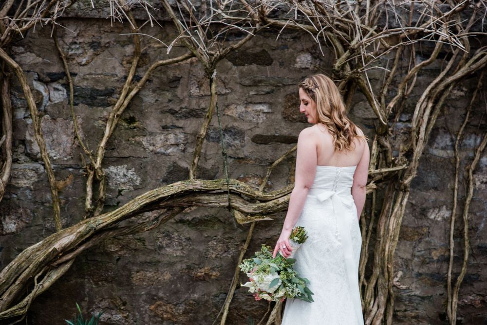 Bride in strapless gown poses beside stone wall with trailing vines.