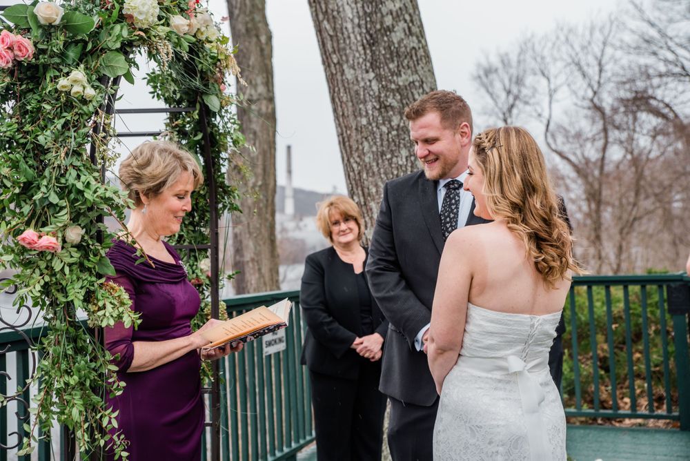 Intimate wedding ceremony on deck with officiant reading from book under floral arch.
