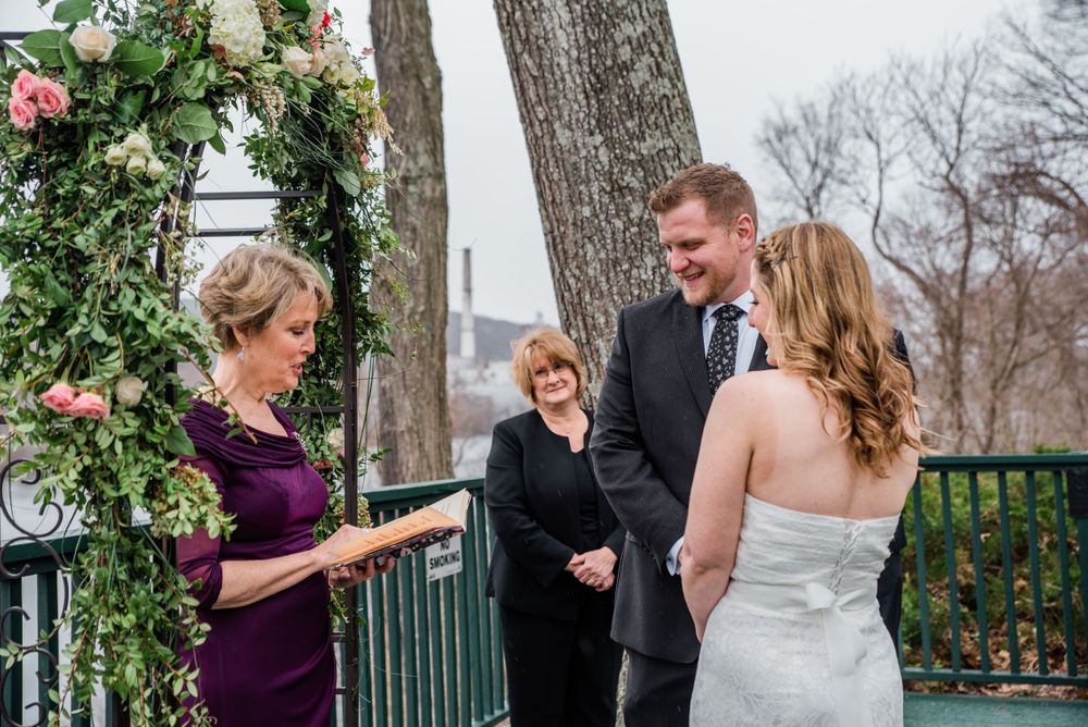 Wedding ceremony takes place on outdoor deck with floral arch and lake view in background.