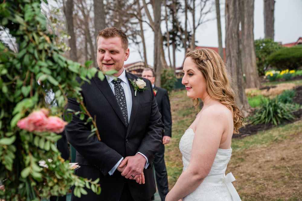 Outdoor wedding ceremony with couple standing beneath flower-adorned archway on wooden deck.