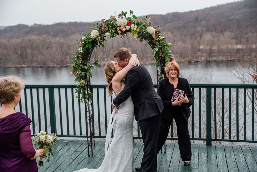 A wedding ceremony takes place on a deck overlooking a lake with a floral arch and winter landscape in the background.