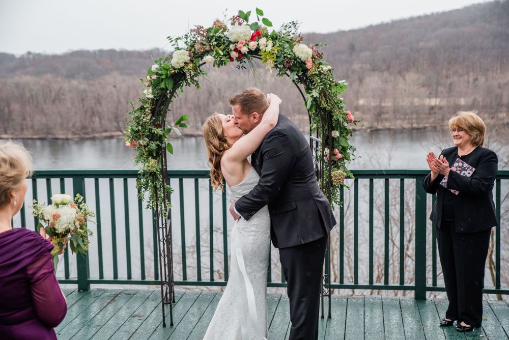 Newlyweds share their first kiss under a floral arch on a lakeside deck during a winter wedding.