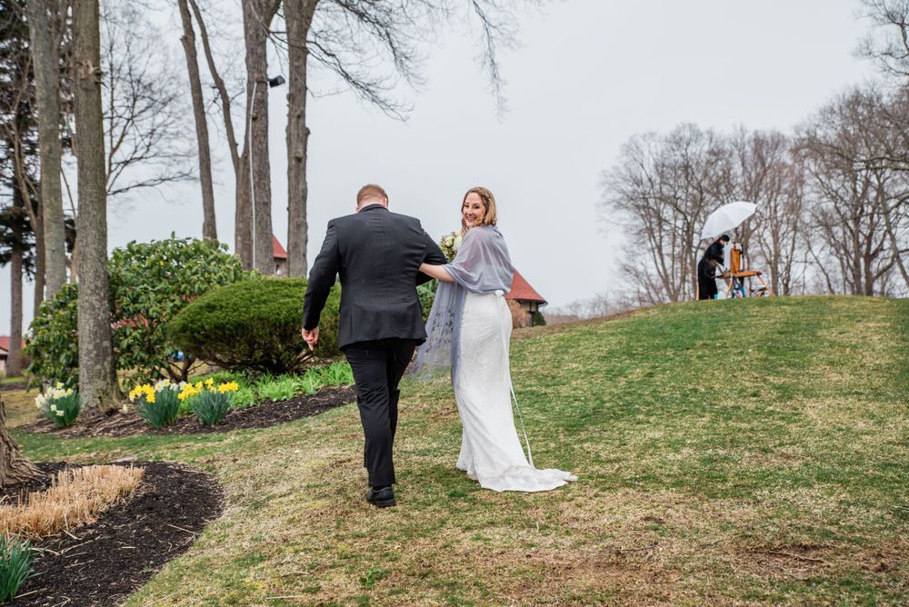 A couple walks together on a grassy hillside with spring daffodils and bare trees in the background.