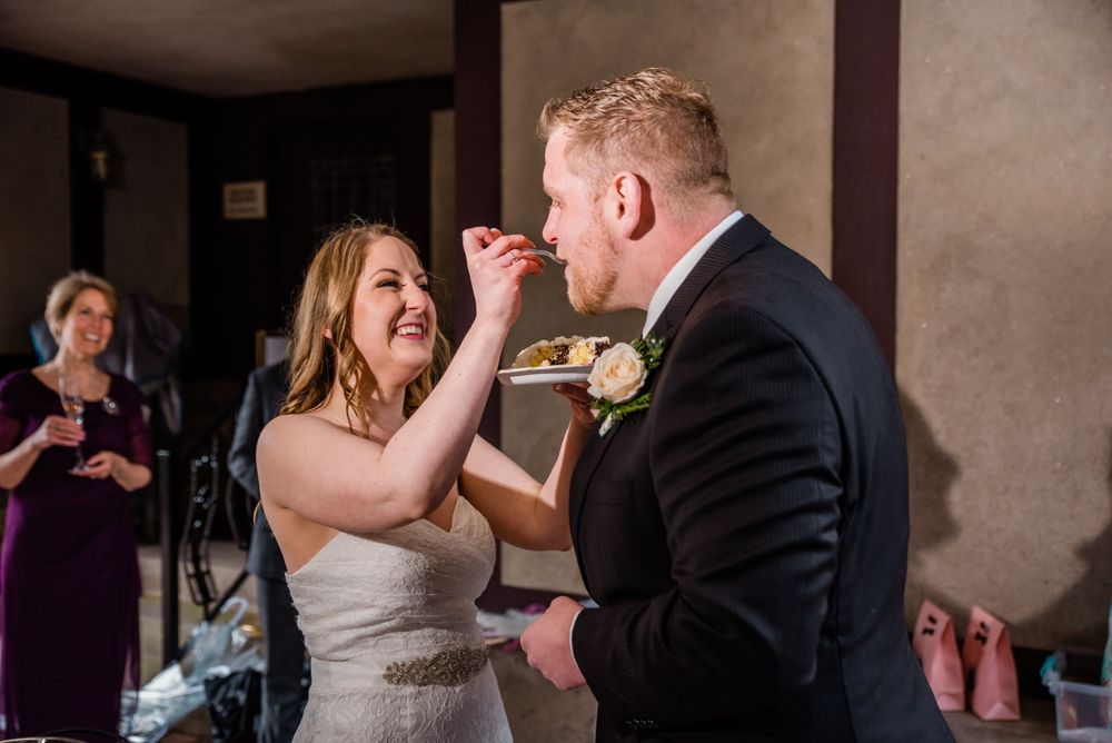Newlyweds feed each other cake in a traditional wedding moment while guests look on.