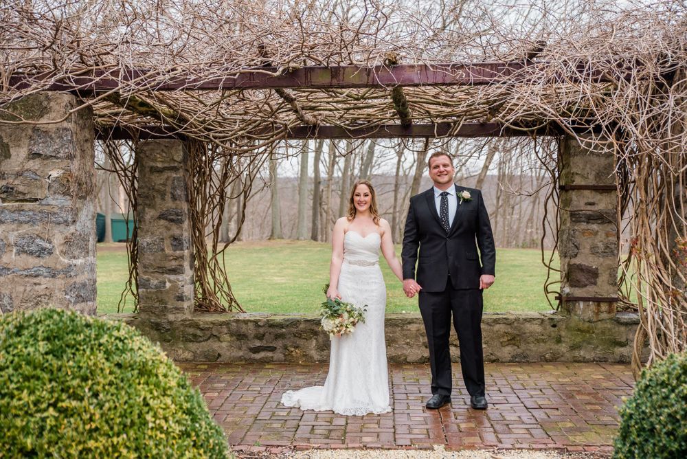A couple poses under a stone archway covered in bare vines in a garden setting.