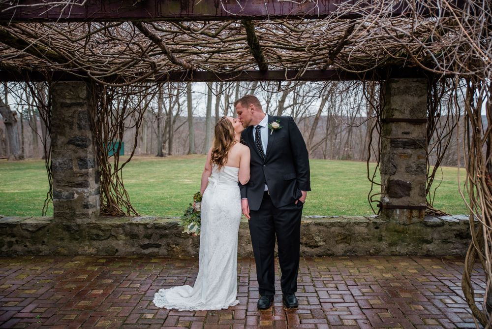 Wedding couple standing beneath a vine-covered stone structure in a garden setting.