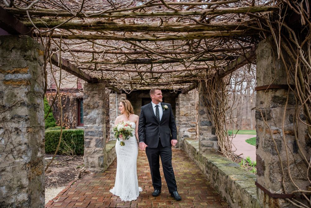 A couple stands in formal attire beneath a stone archway covered in dormant vines.