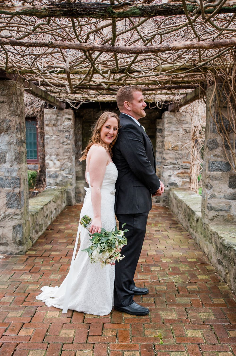 A wedding couple shares a playful moment under a vine-draped stone walkway.