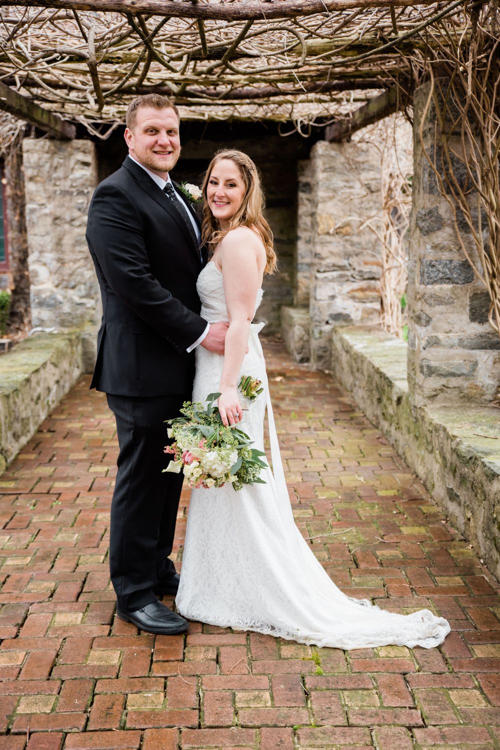 A happy wedding couple poses together under a rustic vine-covered stone archway.