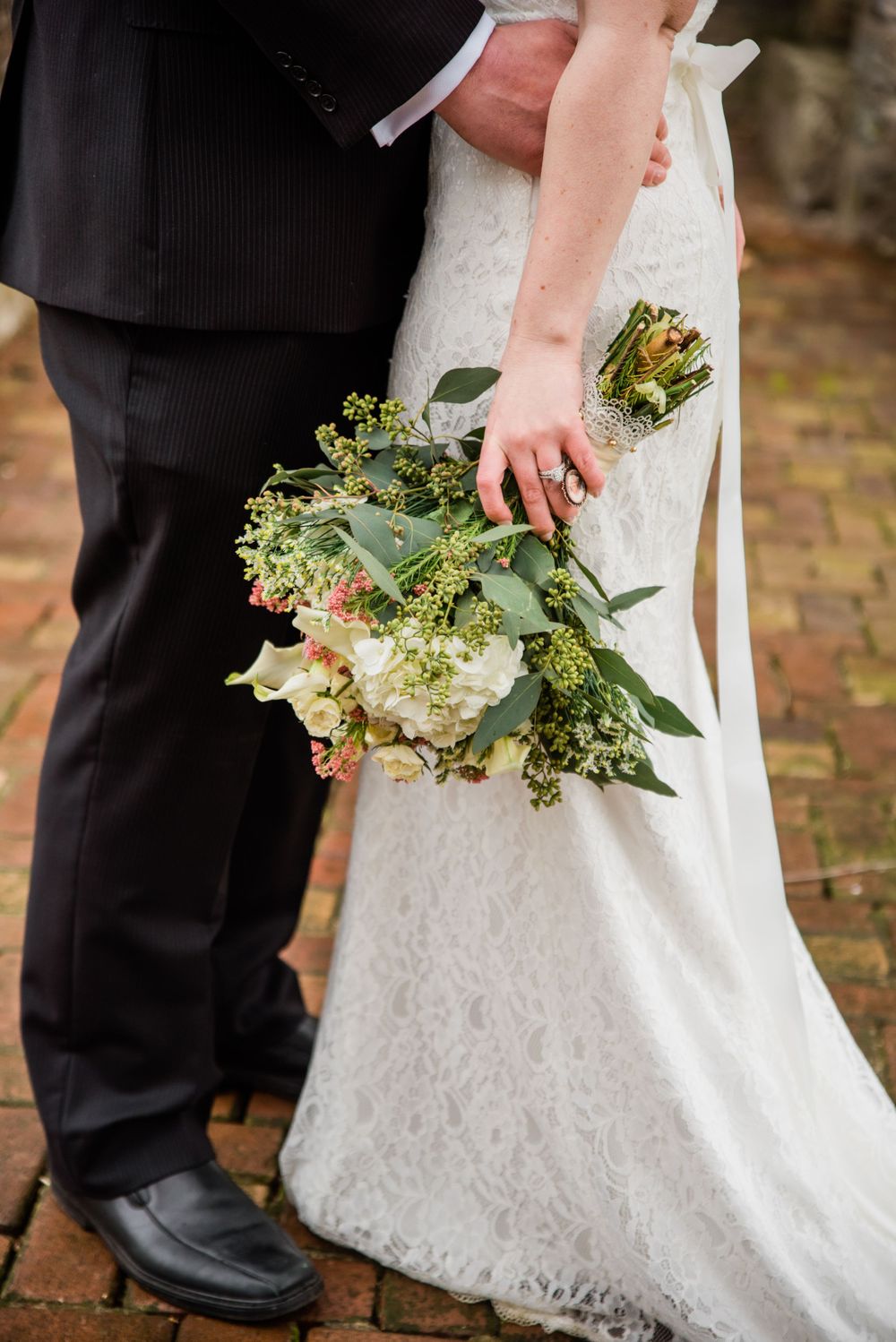Close-up of wedding couple's details with green and white bouquet against white lace dress.