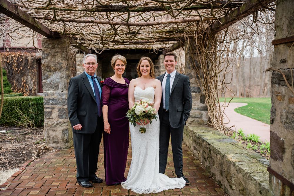 Family wedding portrait under stone pergola with vines and brick pathway.