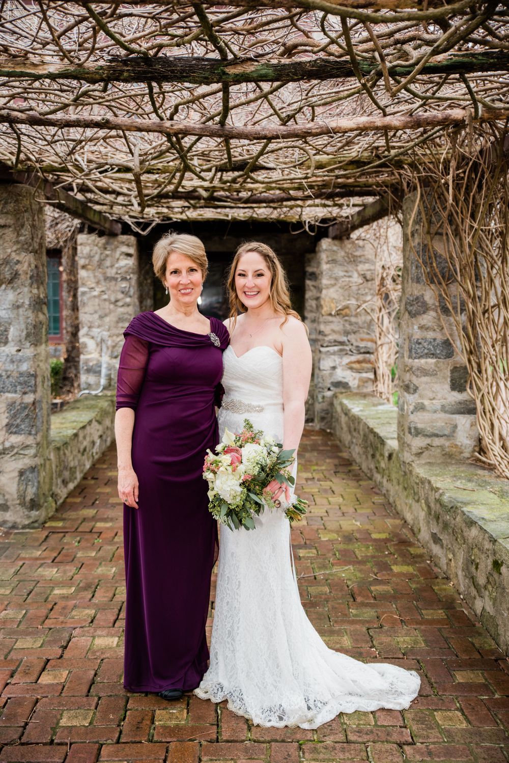 Formal wedding portrait of two women in purple and white dresses under stone archway.