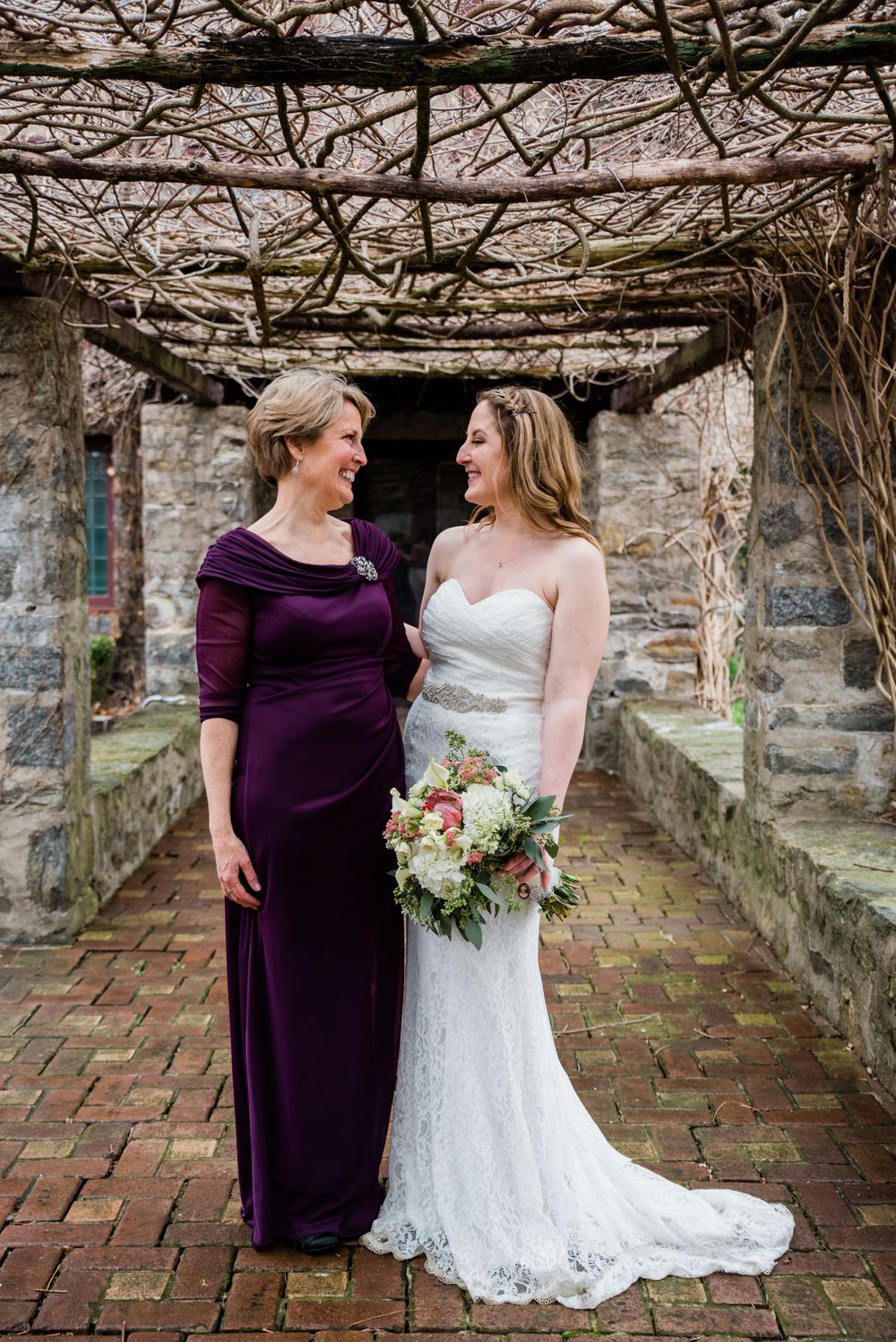 Two women share an intimate moment under vine-covered stone pergola at wedding.