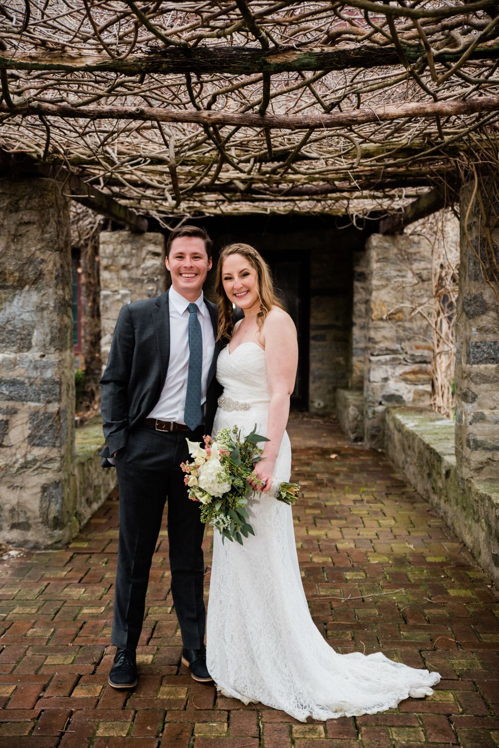 A couple poses in formal wedding attire under a stone archway covered in bare vines and branches.