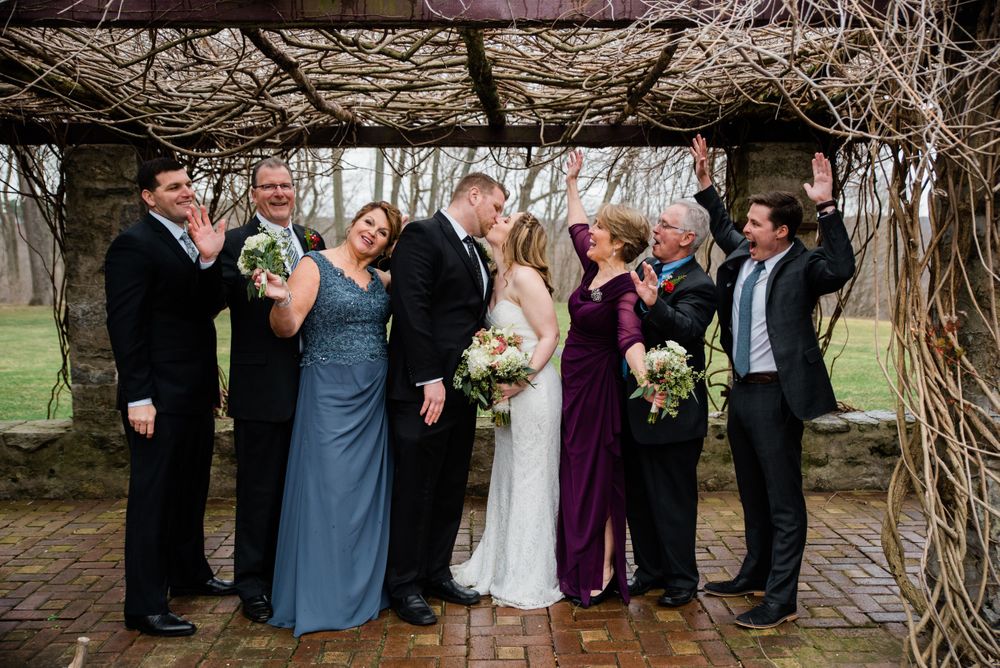 Wedding party laughs together while posing beneath a vine-covered stone pergola.