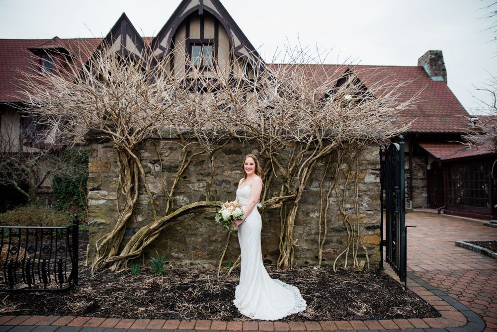 Bride poses in fitted white gown against stone wall and bare winter trees.