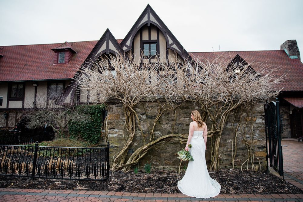 White wedding dress contrasts against stone Tudor-style building with bare winter branches.