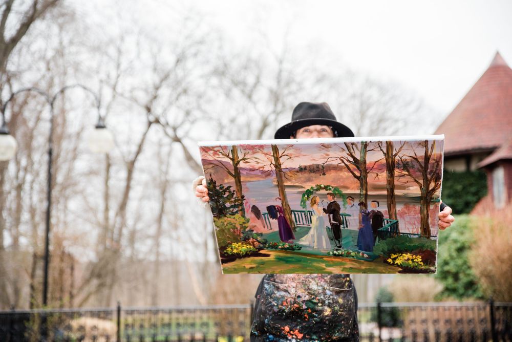 Person holds up colorful landscape painting in front of winter garden scene.